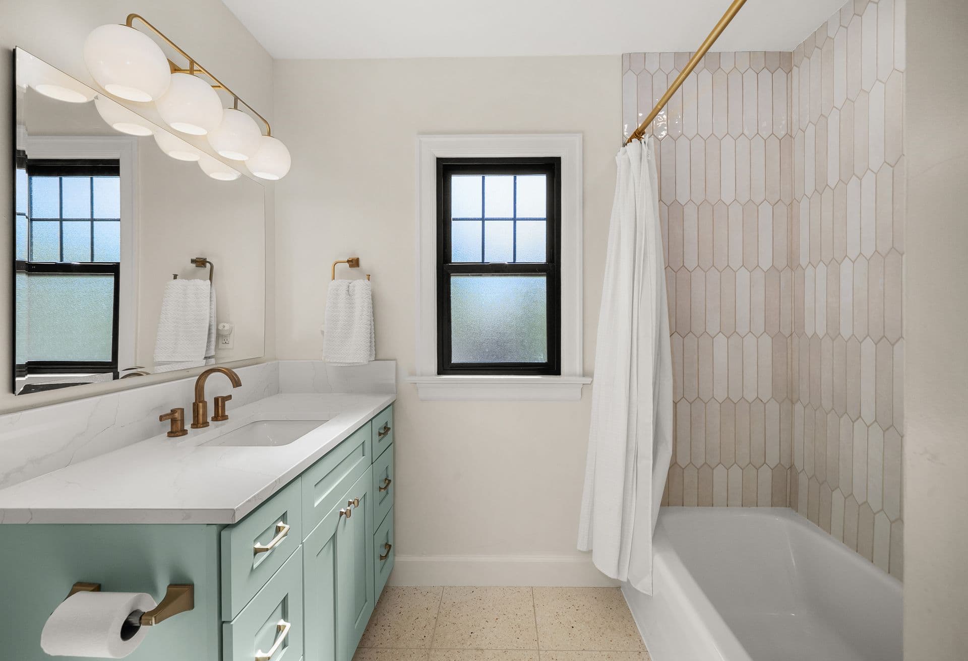 Modern bathroom with mint-green vanity, marble countertop, brass fixtures, hexagonal tile shower surround, and frosted windows