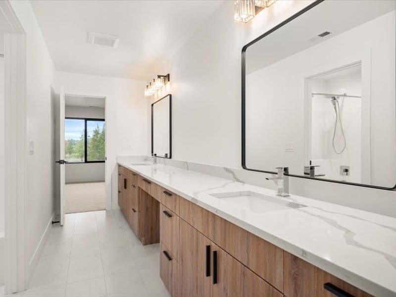 A white bathroom with long double vanity with white and gray marbled countertop, two large mirrors, and brown cabinets.